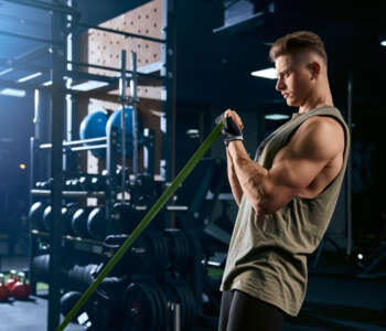 Portrait of muscular bodybuilder training arms with fitness resistance bands. Side view of man with perfect body training biceps in gym in dark atmosphere. Concept of bodybuilding.